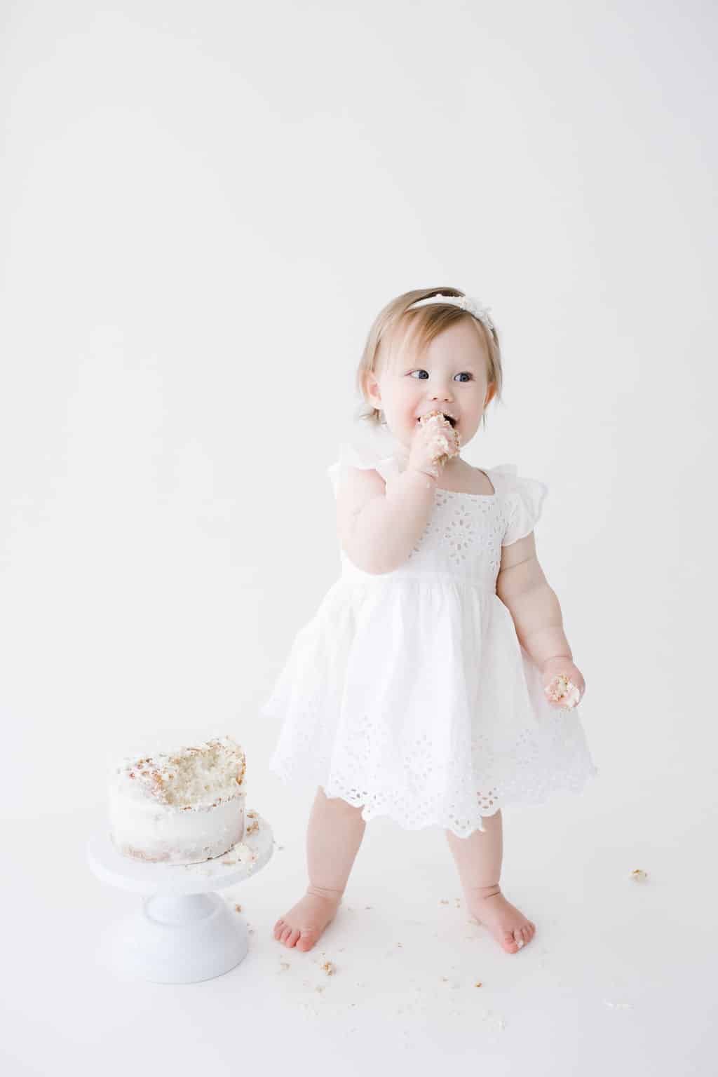 A toddler in a white dress stands barefoot on a white floor, eating cake with one hand. A partially eaten cake sits on a stand nearby, with crumbs scattered around.