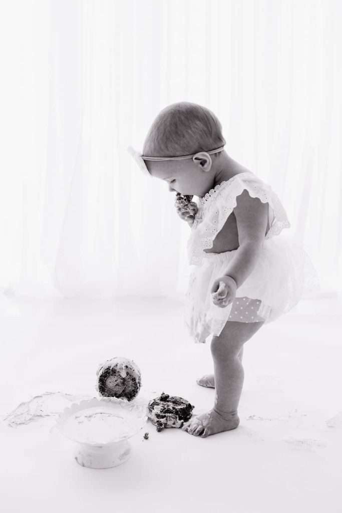 Baby in a dress stands on a white floor, eating cake with hands. A bowl and messy cake are on the ground. Light background.