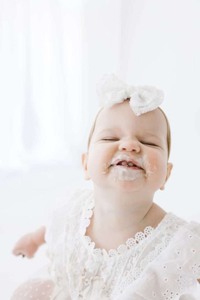 Smiling baby in a white dress and bow has food smeared around their mouth. The background is light and softly blurred.