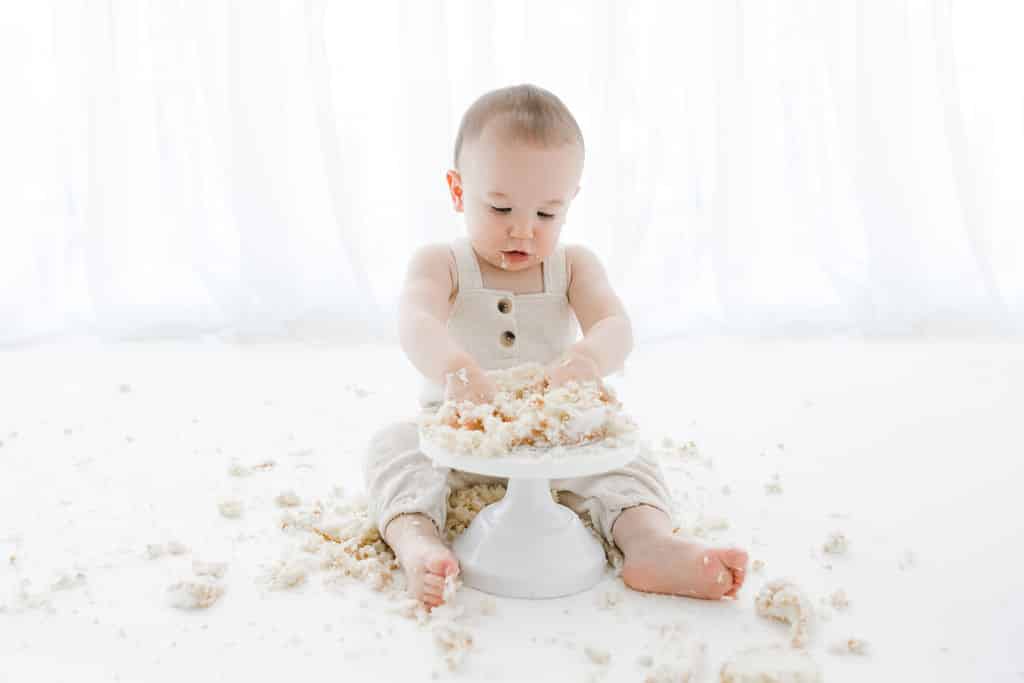 A baby in light overalls sits on the floor, playing with and smashing a white cake on a stand. Cake crumbs are scattered around on a white background.