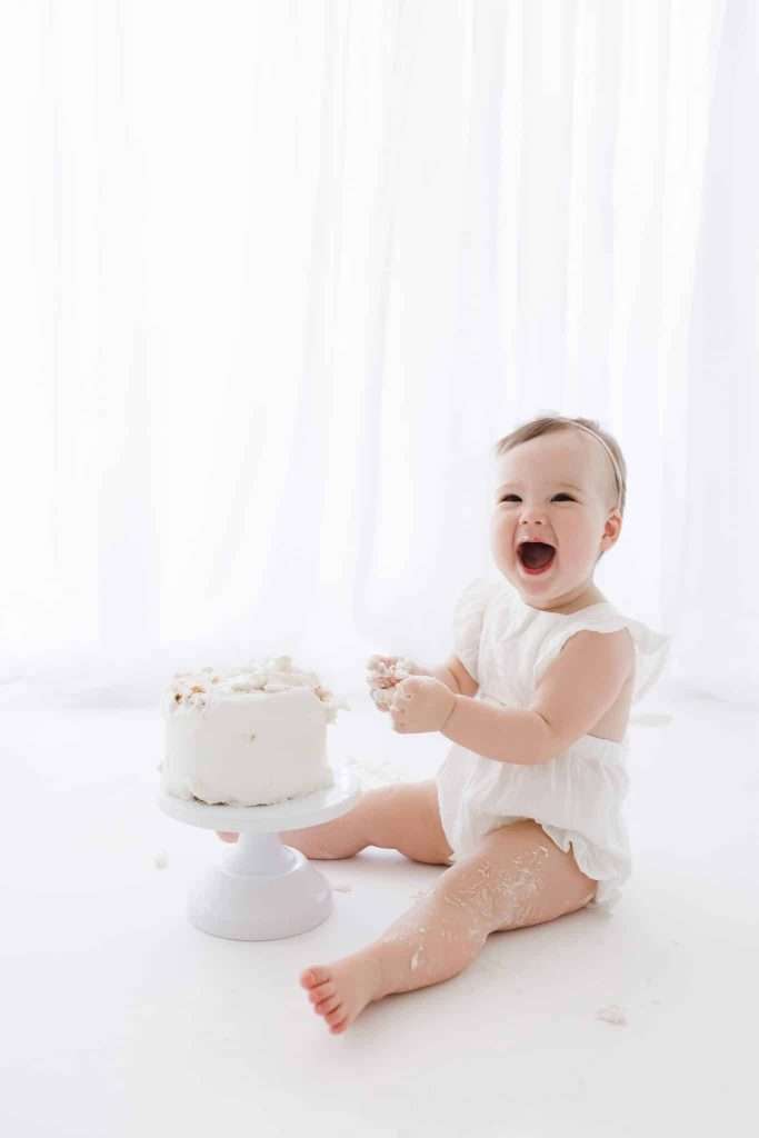 A baby in a white outfit sits on the floor next to a white frosted cake, smiling with hands covered in cake, in front of a sheer white curtain.