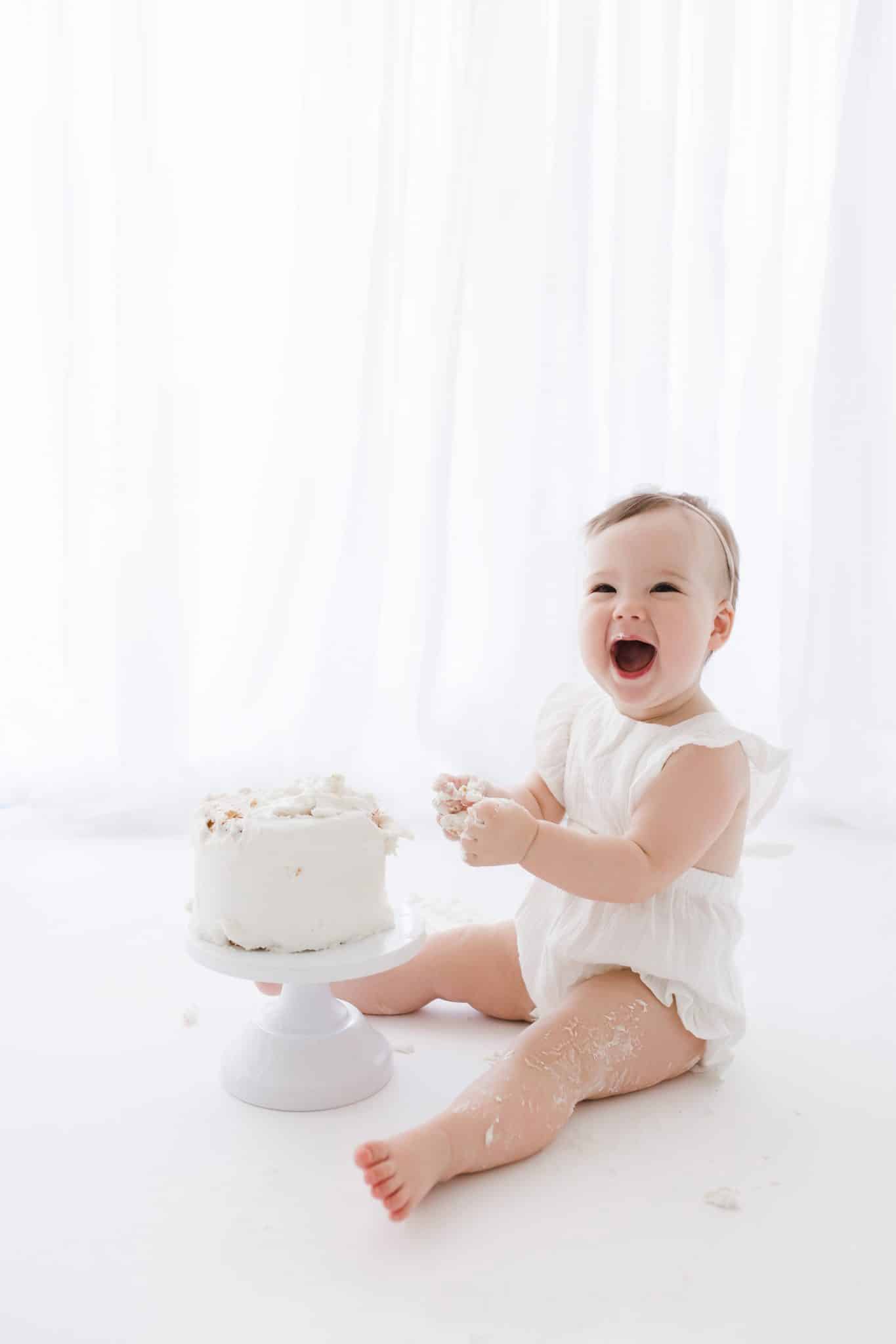 A baby in a white outfit sits on the floor next to a white frosted cake, smiling with hands covered in cake, in front of a sheer white curtain.