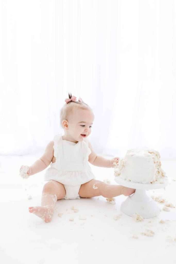 A baby in a white outfit sits on the floor, smiling and playing with a partially smashed cake on a stand.