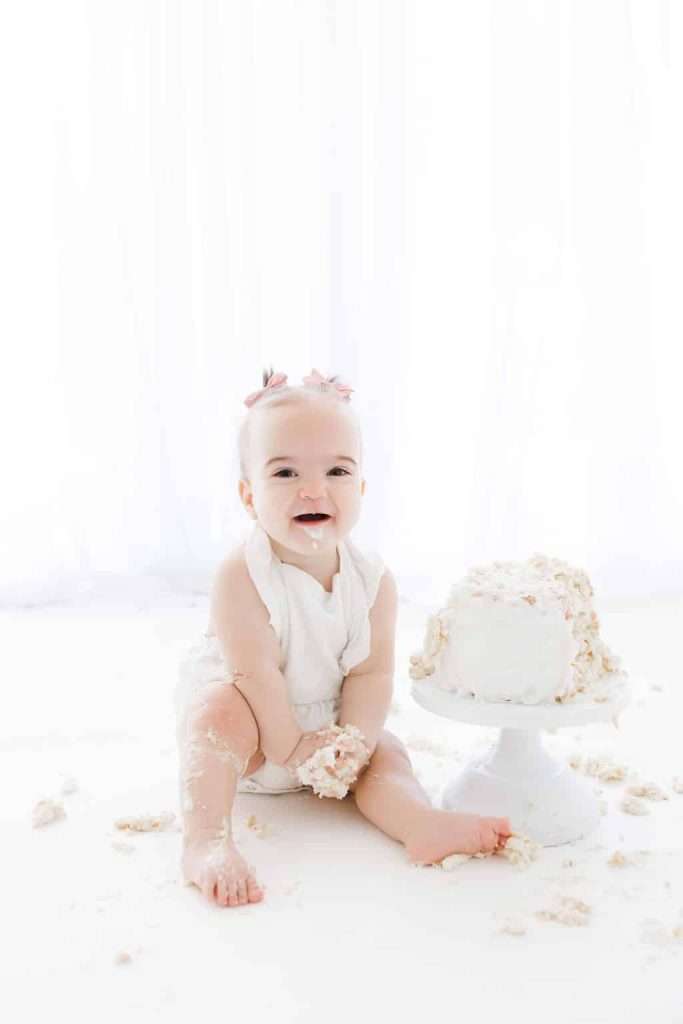 Baby in a white outfit sits on the floor, smiling, with hands and feet covered in cake beside a partially smashed white cake on a stand.