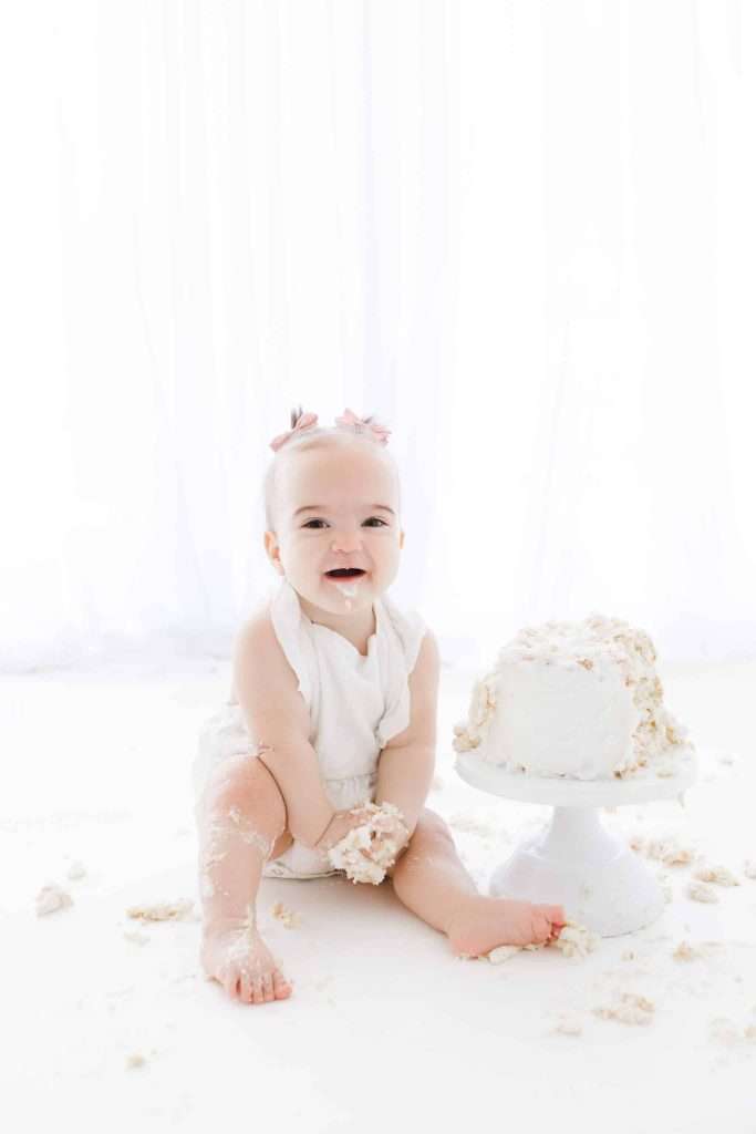 A baby in a white outfit sits on the floor with a smashed white cake on a stand, smiling with cake and frosting on hands and legs.