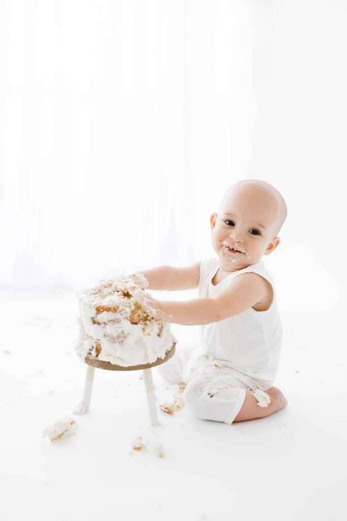 A baby in white clothes sits on the floor, smiling with cake on their face and hands, while smashing a small cake on a stand.