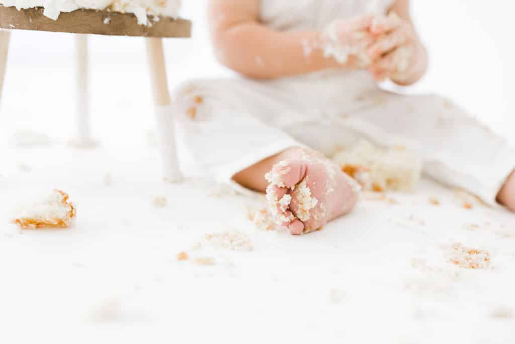A baby in white clothing sits on the floor with cake crumbs scattered around, holding cake in one hand and some cake on their foot.