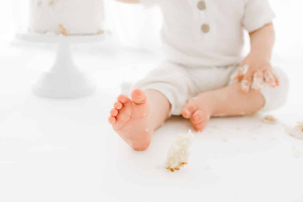 A baby in white clothes sits on the floor with cake crumbs around, one foot in focus and a partially eaten cake in the background.