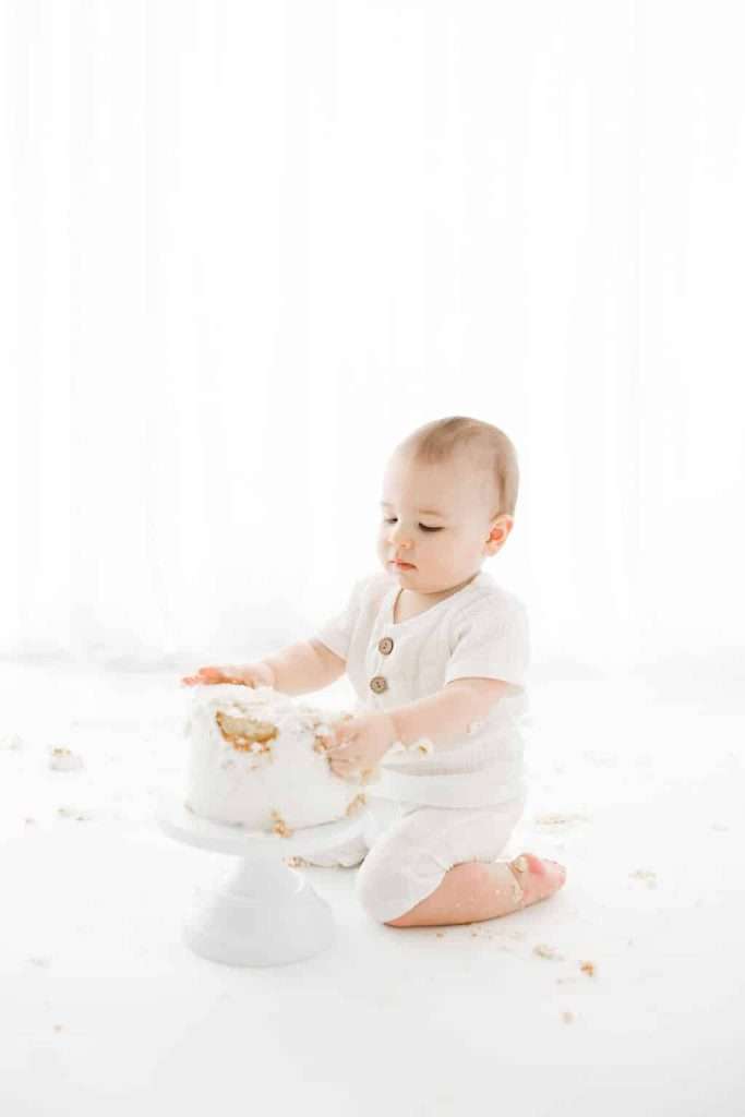 Baby in white outfit sits on the floor, touching a partially eaten cake on a stand, with cake crumbs scattered around.