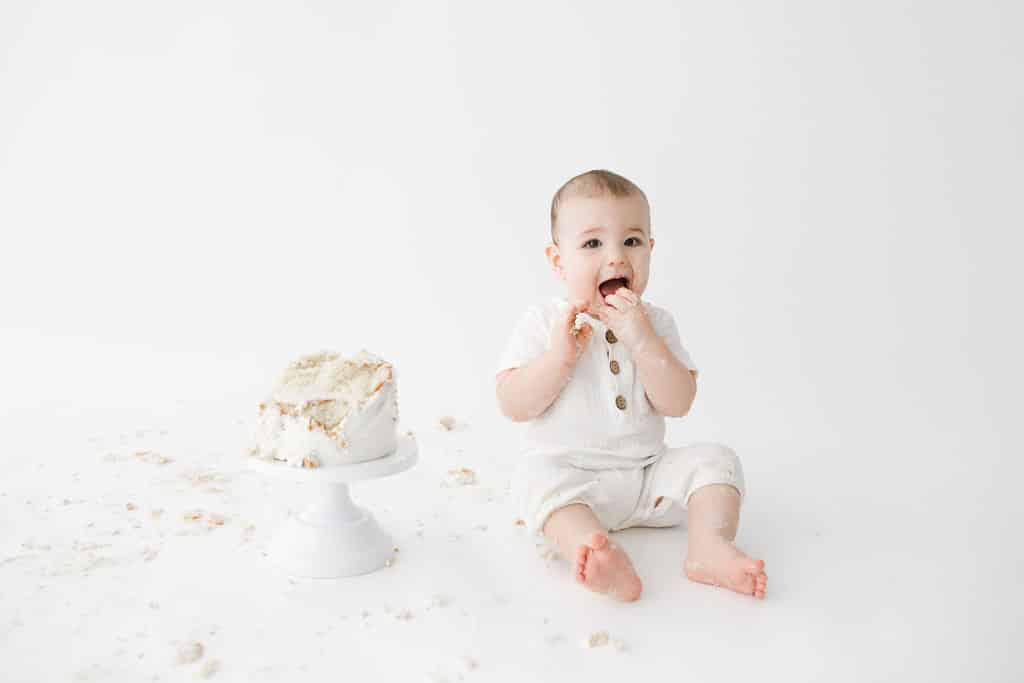 A baby in a white outfit sits on the floor next to a partly eaten cake, eating with hands. Cake crumbs are scattered around on a white background.