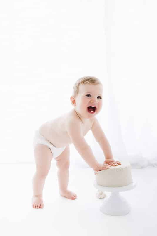 A baby in a diaper stands and leans on a white frosted cake placed on a cake stand, smiling in a bright, white setting.
