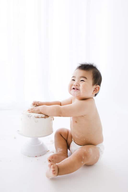 A baby in a diaper sits on the floor, smiling and playing with a frosted cake on a cake stand in a bright, white setting.