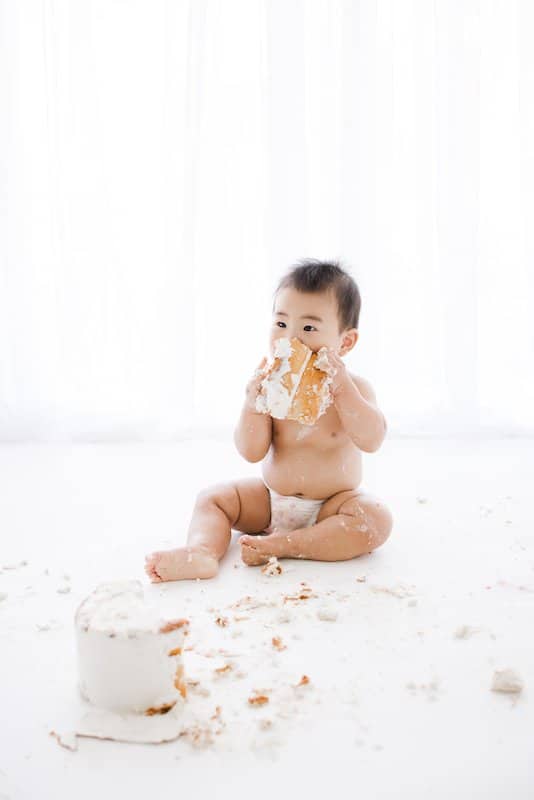 A baby in a diaper sits on the floor, holding and eating a piece of cake, with cake and crumbs scattered around.