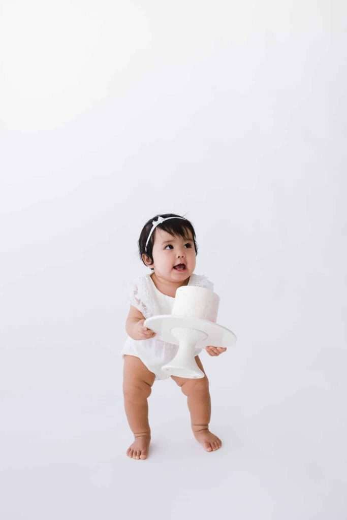 A baby in a white outfit and headband holds a white cake stand with a small cake against a plain white background.