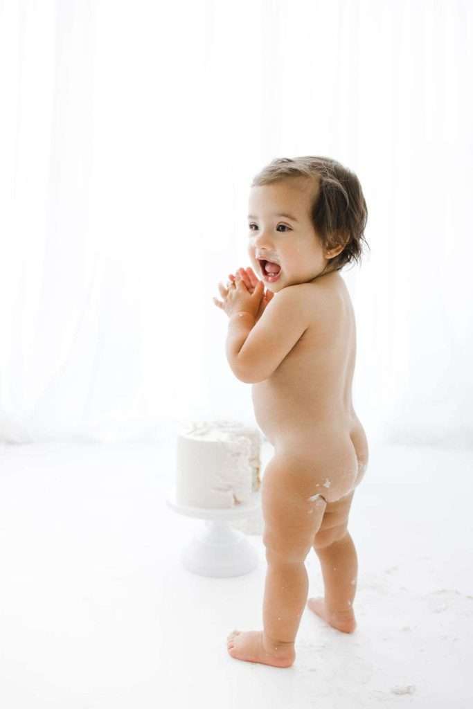 A baby stands naked on a white floor next to a small white cake, looking over their shoulder and smiling.