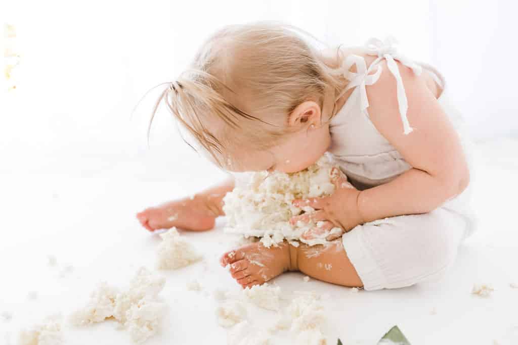 Baby in white outfit sits on floor, holding and eating a messy piece of cake with both hands. Cake crumbs and frosting are scattered around.
