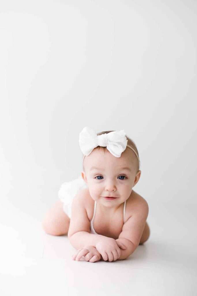 Baby with a white bow headband lies on her stomach on a white background, looking at the camera with a slight smile.