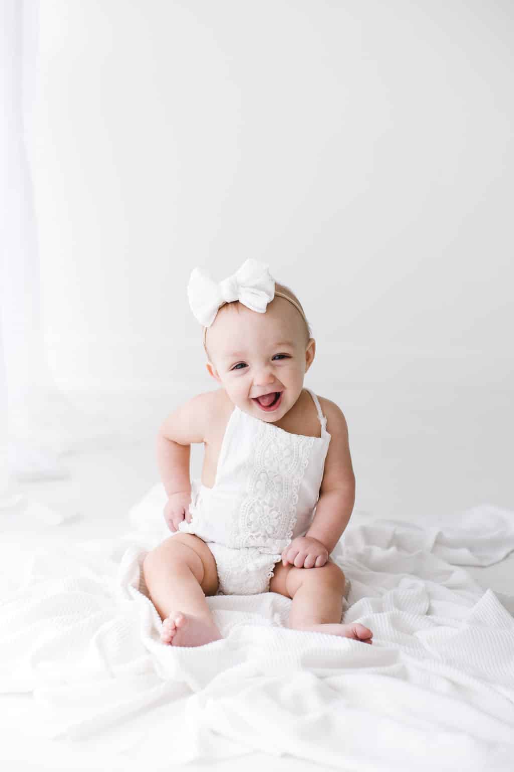 Smiling baby wearing a white outfit and large white bow sits on a white blanket against a plain white background.