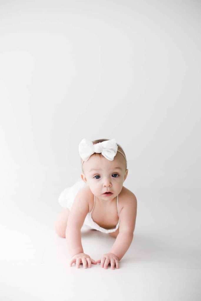Baby wearing a white bow headband and white outfit crawls on a white surface, facing the camera, with a neutral background.