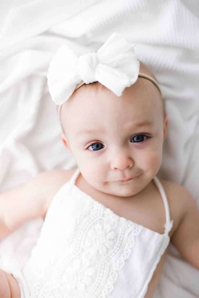 Baby wearing a white outfit and a large white bow headband, lying on a white blanket, looking up at the camera.