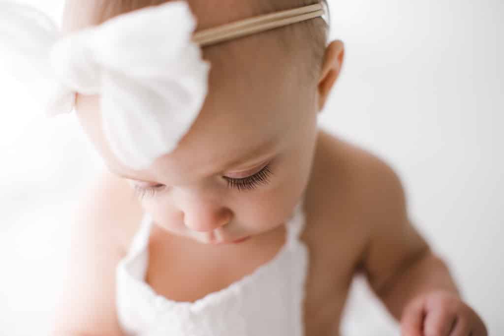 Baby wearing a white outfit and large white bow headband looks down, showing long eyelashes and soft focus background.