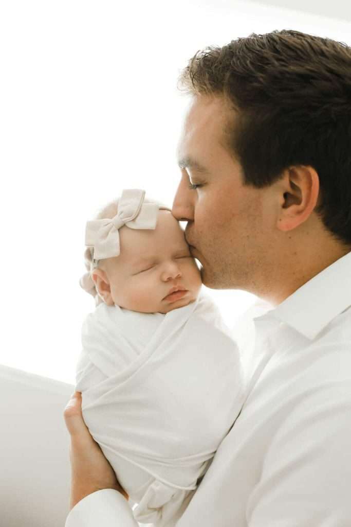 Man in a white shirt holds and kisses a sleeping baby wrapped in a white blanket with a bow headband.