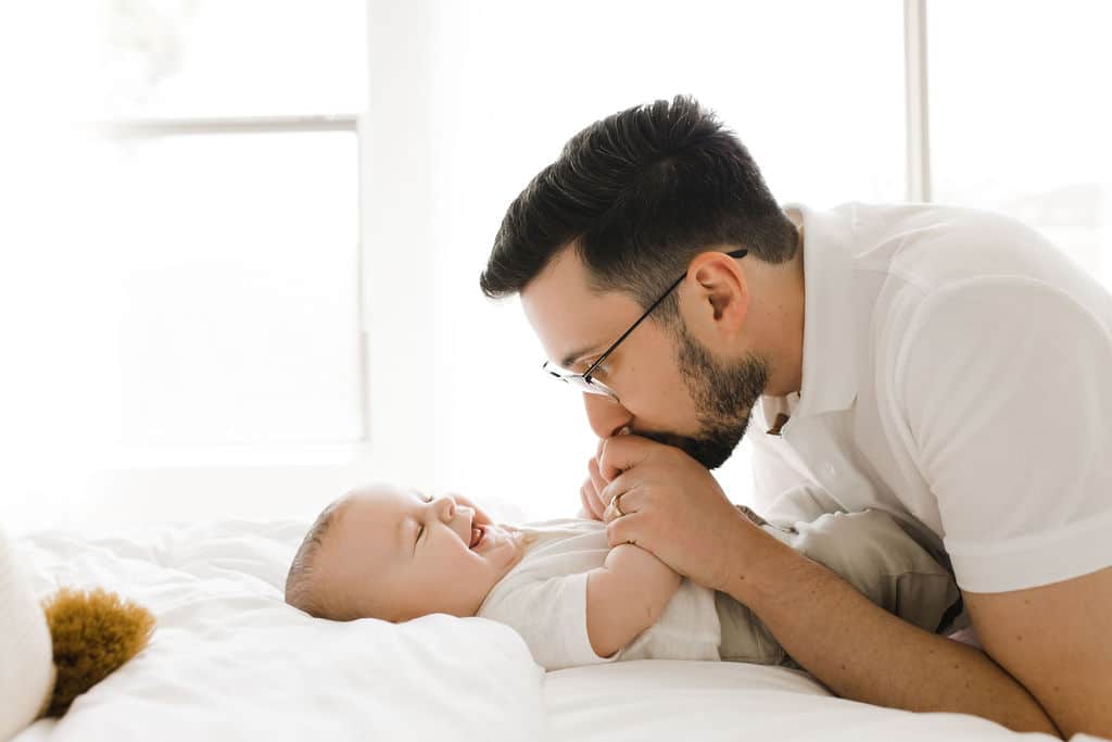 A man lying on a bed kisses a smiling baby’s feet. They are both dressed in light-colored clothes in a bright room.
