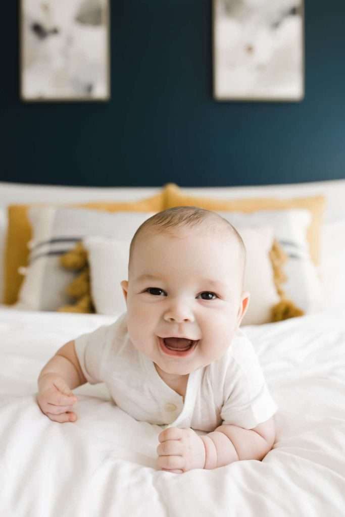 Smiling baby lying on a bed in a white onesie, with decorative pillows and two paintings visible in the background.