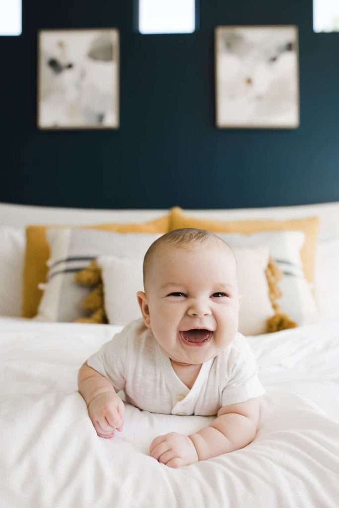 Smiling baby lying on a bed with white linens and decorative pillows, with abstract art and a dark wall in the background.