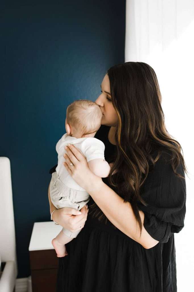 Woman in a black dress holds and kisses a baby on the head in a room with a dark blue wall and a window with white curtains.