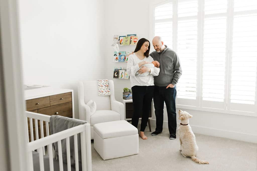 A woman and man stand in a bright nursery holding a newborn, while a dog sits on the carpet looking up at them.
