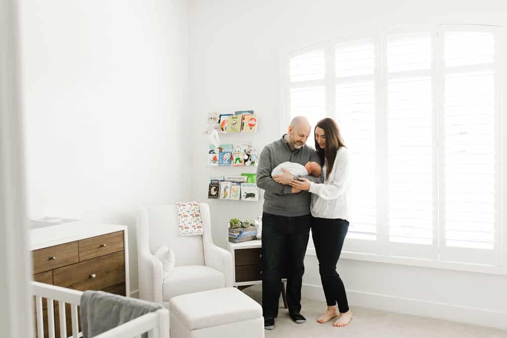A couple stands in a bright nursery, holding a newborn baby. There is a crib, chair, dresser, and shelves with books in the background.