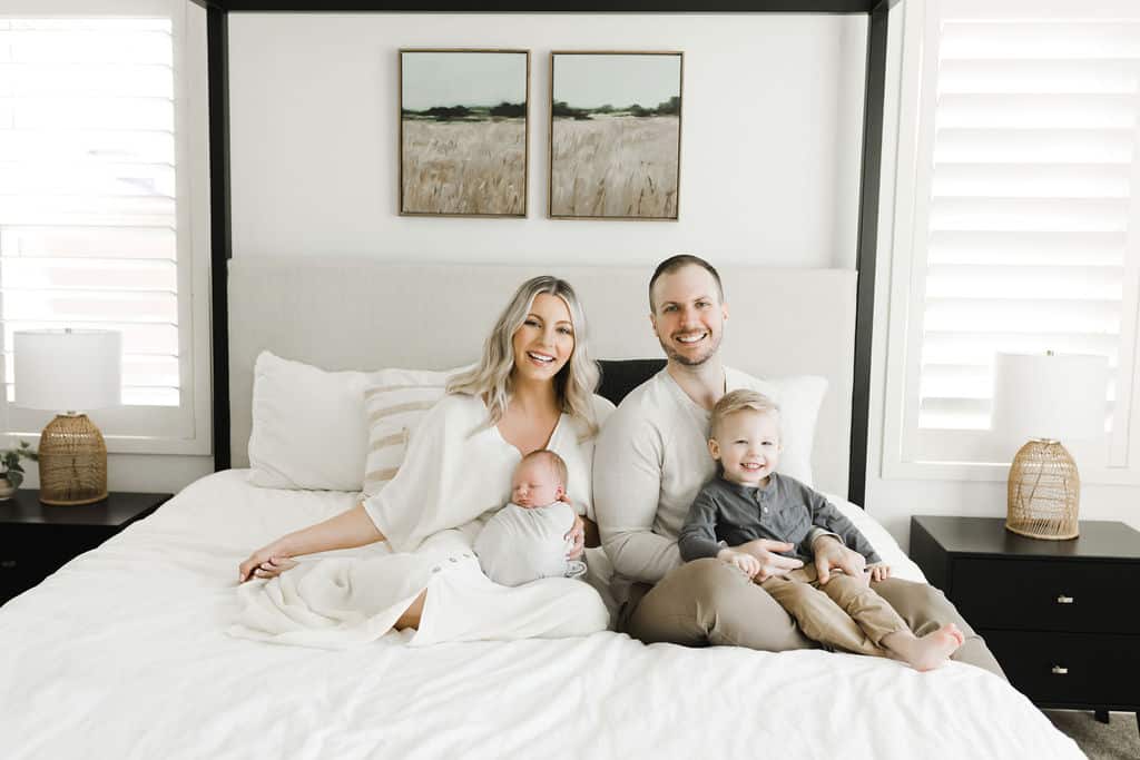 A family of four sits on a bed. Two adults sit in the center, holding a baby and a young child. The room has white walls, a headboard, and two bedside tables with lamps.