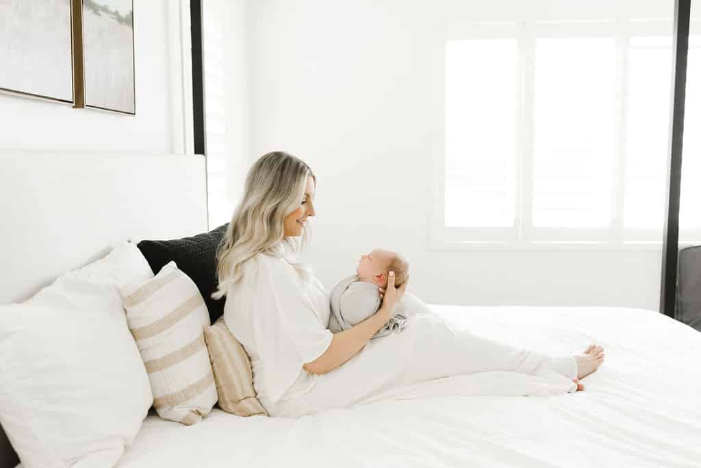 Woman sits on a white bed, holding a baby in her lap. The room is bright with natural light from a nearby window.