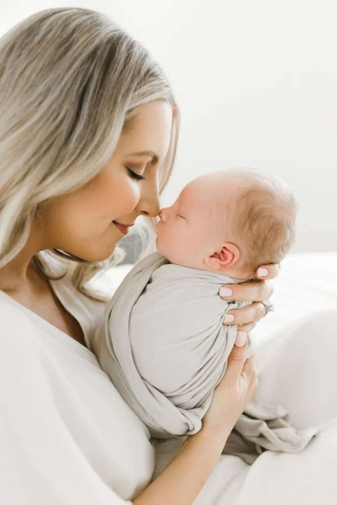 Woman with long, blonde hair gently holds and touches noses with a swaddled newborn baby, both wearing light-colored clothing in a bright setting.