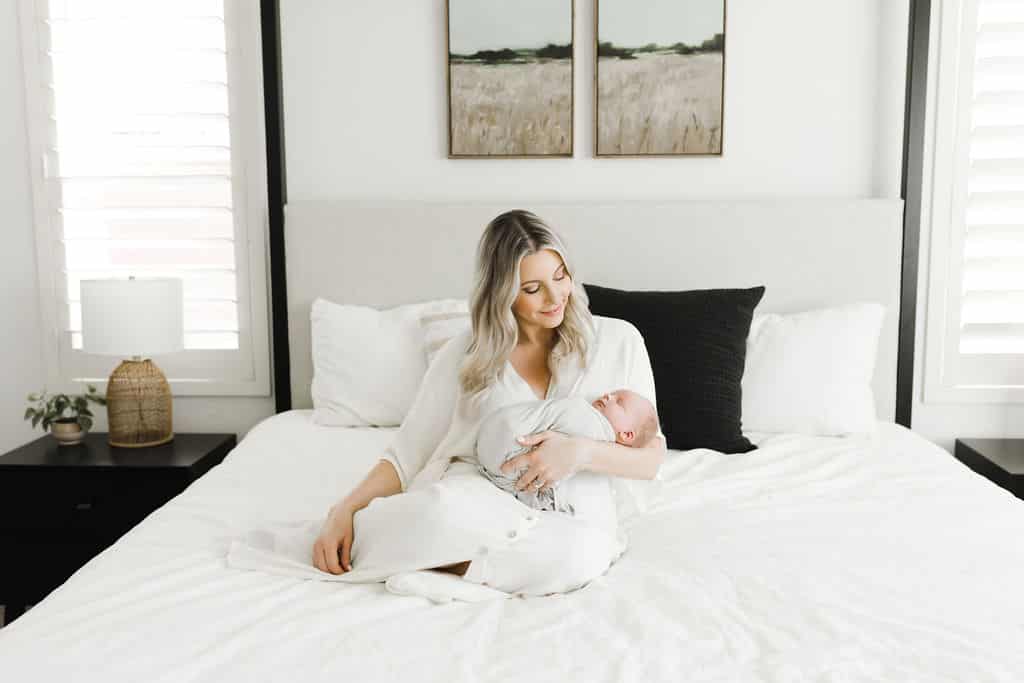 Woman in white robe sits on a bed, holding a swaddled baby in her arms. The room is bright with white bedding, two paintings above the bed, and a lamp and plant on nightstands.