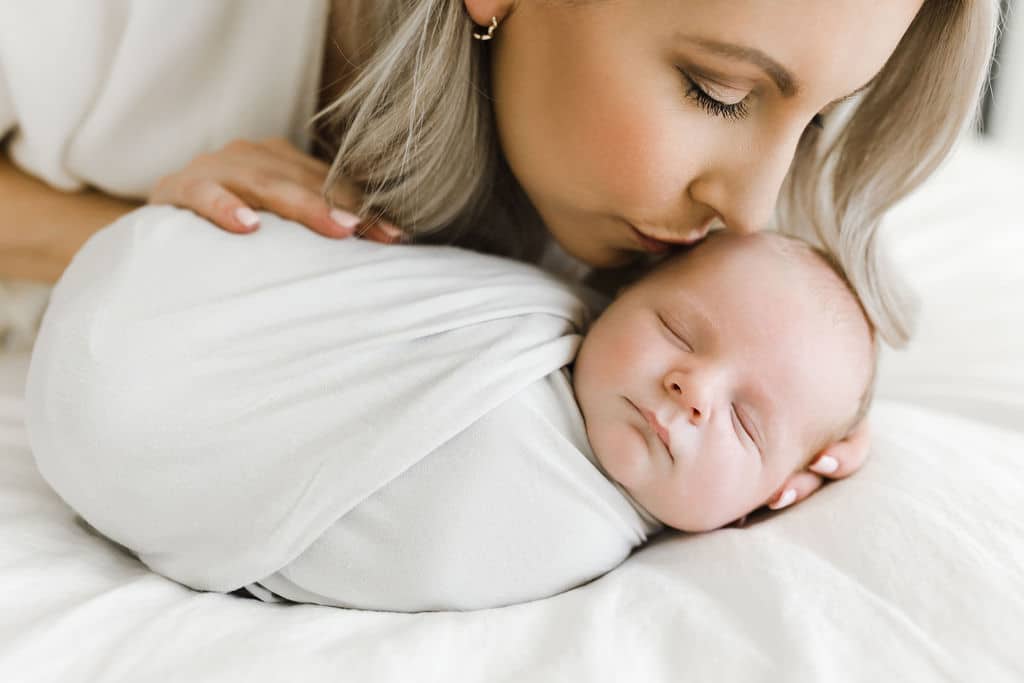 Woman with blonde hair kisses the forehead of a sleeping newborn wrapped in a white blanket, both lying on a white bed.