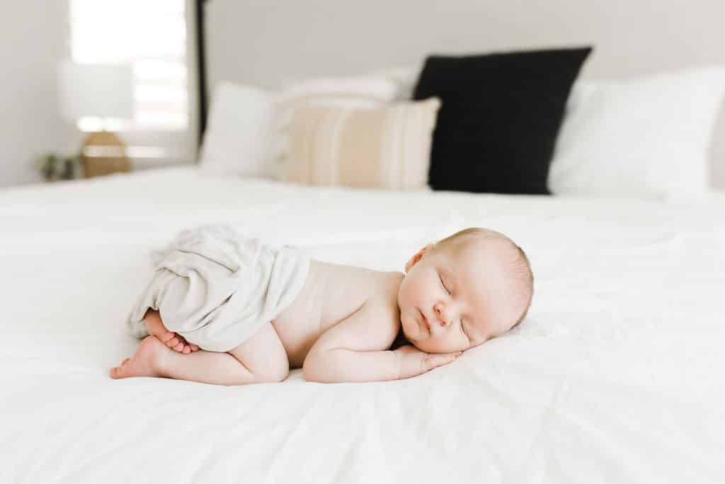 Newborn baby sleeping on a white bed, partially covered with a light blanket, with pillows and a lamp visible in the background.