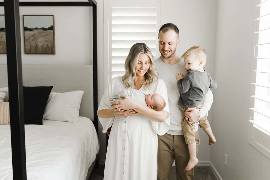 A woman holds a newborn while a man stands beside her holding a toddler in a bright bedroom with a bed and window shutters.