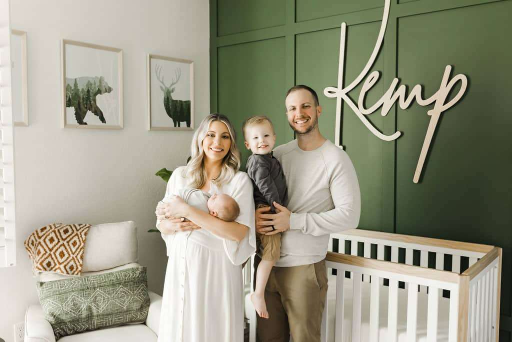 A family of four stands in a nursery with green walls, a crib, and animal artwork. The mother holds a baby, the father holds a toddler, and "Kemp" is written on the wall behind them.