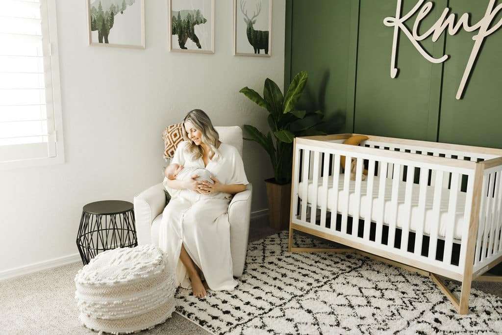 A woman in a white robe sits in a nursery, holding a baby. There is a white crib, green accent wall, plant, round ottoman, and wall art with animal prints in the room.