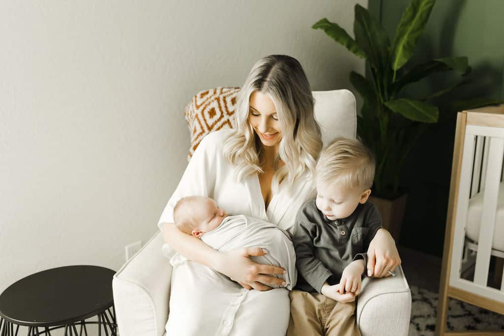 A woman sits in a white chair holding a newborn baby, with a young child sitting beside her and a green plant in the background.