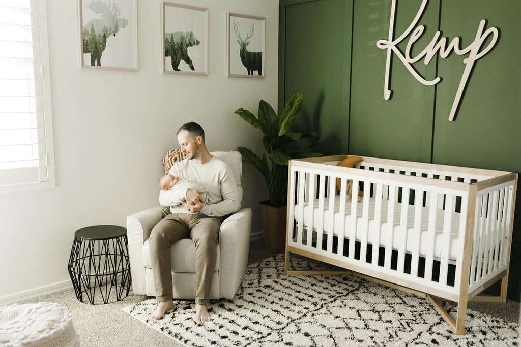 A man sits in a nursery holding a baby. The room features a white crib, green accent wall, framed animal art, and a sign reading “Kemp.” There is a plant and patterned rug on the floor.