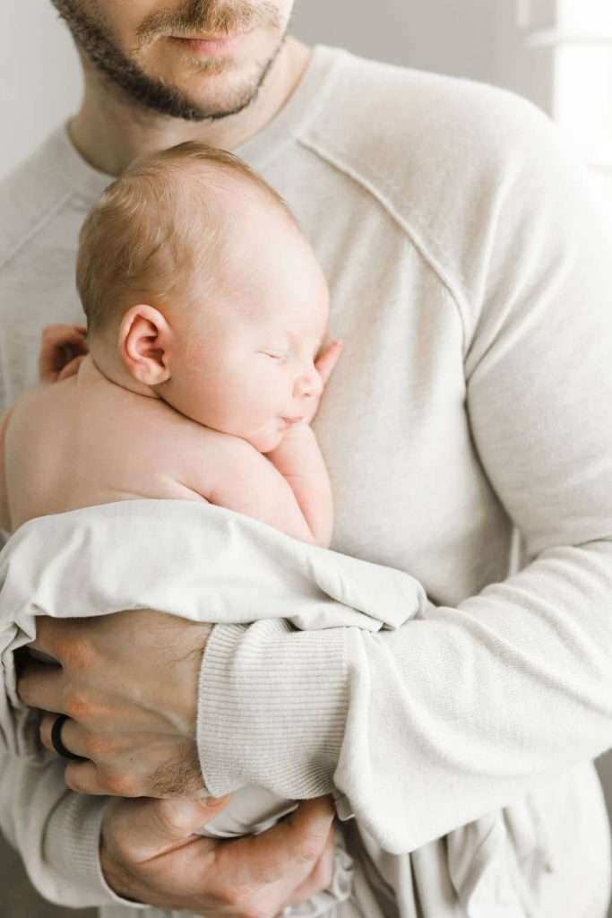 An adult holds a sleeping newborn baby wrapped in a light blanket against their chest. Both are dressed in light-colored clothing. The adult's face is partially visible.