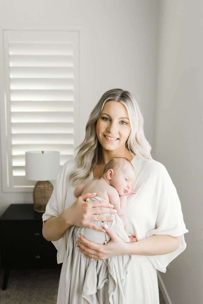 Woman with long blonde hair holds a sleeping baby wrapped in a blanket in a bright room with a window, lamp, and black nightstand.