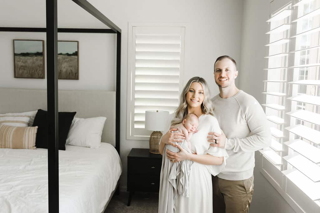 A couple stands in a bright bedroom, smiling at the camera while the woman holds a sleeping newborn wrapped in a blanket.