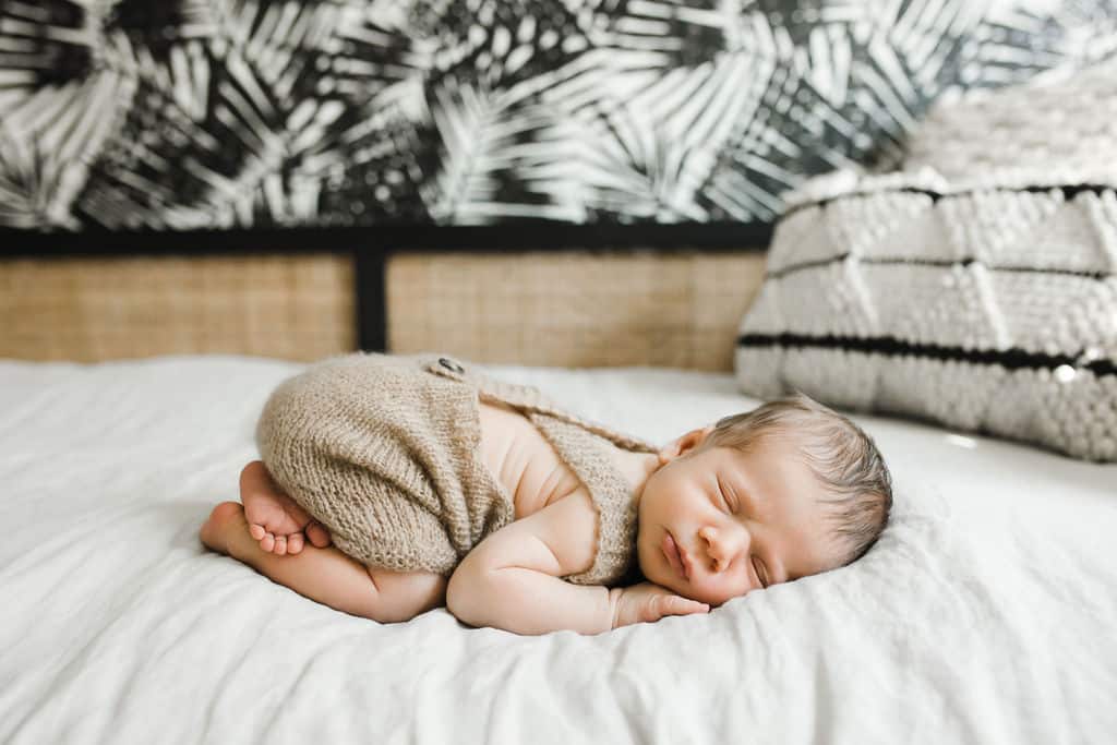 Newborn baby wearing a knitted outfit sleeps curled up on a white bed with a decorative black and white pillow and patterned wall in the background.