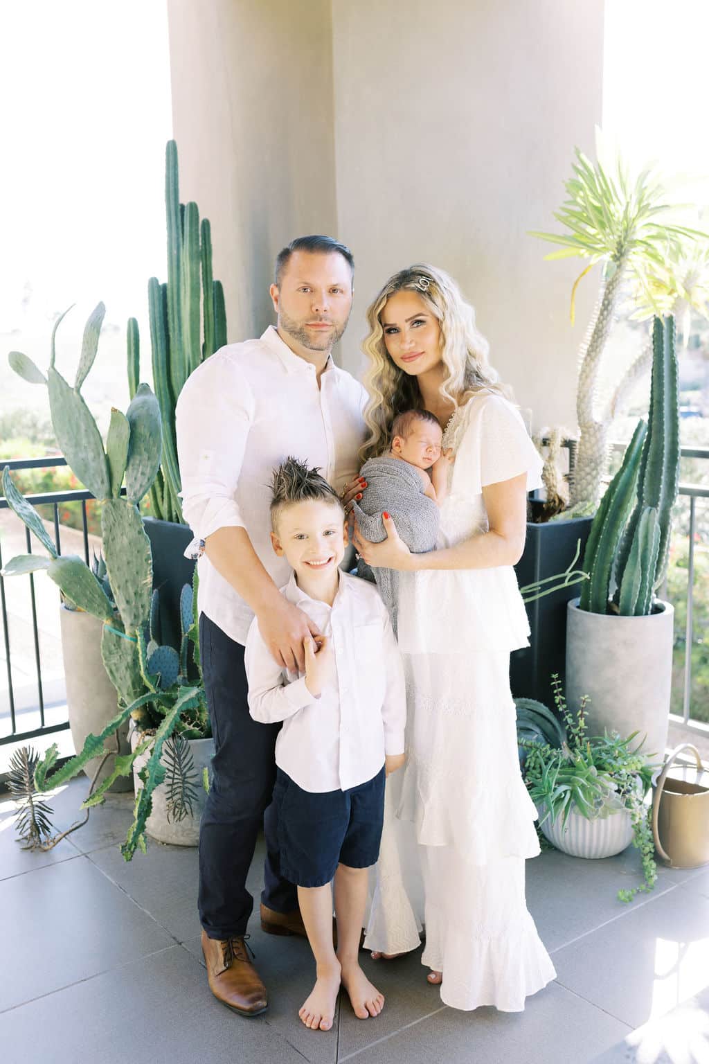 A family of four stands on a patio with potted plants, the father and mother each holding a child, all dressed in white and light-colored clothing.