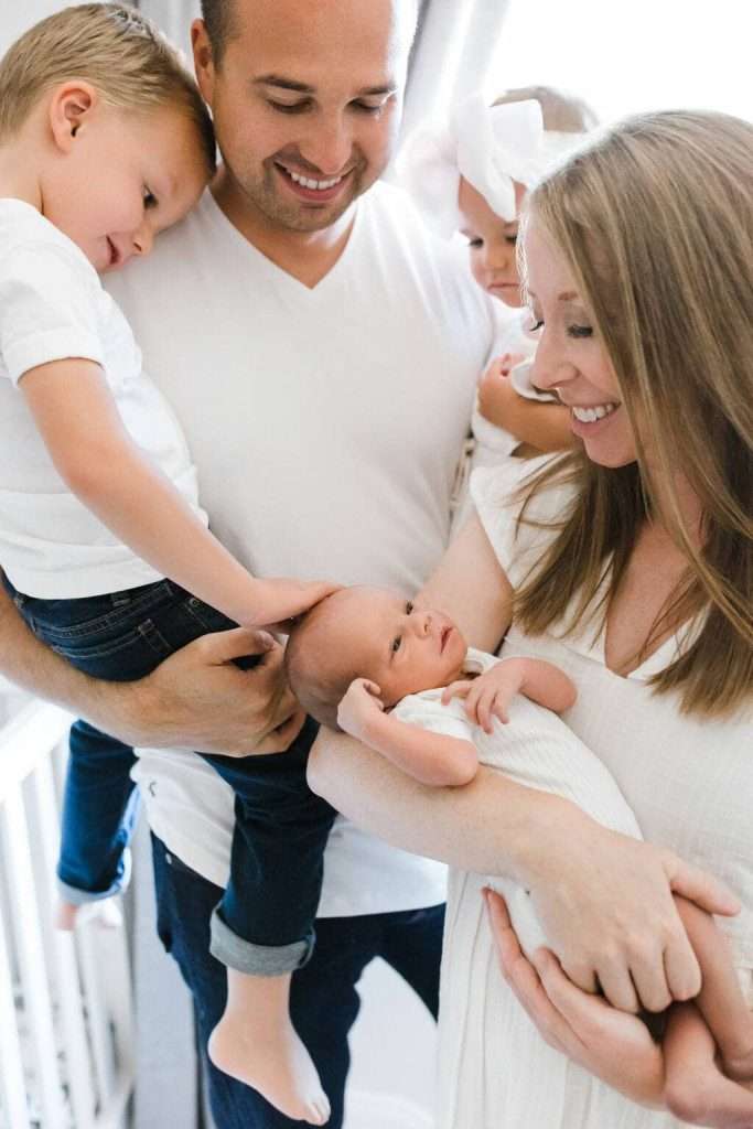 A smiling family of four, with parents holding a newborn and toddler, while an older child gently touches the baby's head.