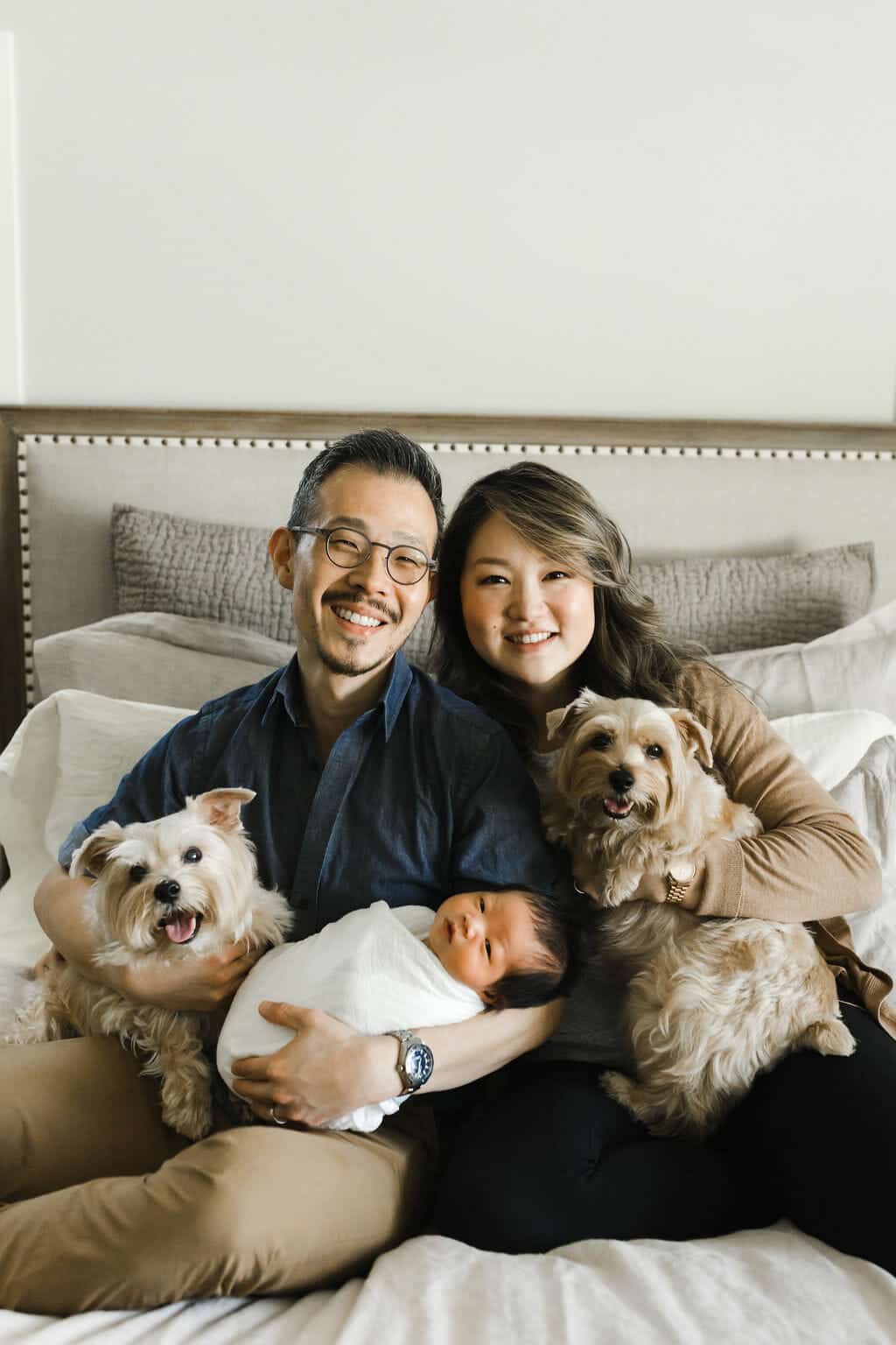 A couple sits on a bed, holding a newborn baby wrapped in a white blanket, with two small dogs on their laps.